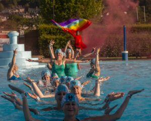 line of artistic swimmers in water with indian dancer dressed in red behind with red smoke and pride flag