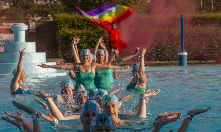 line of artistic swimmers in water with indian dancer dressed in red behind with red smoke and pride flag