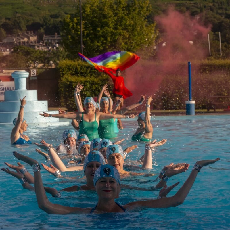 Pride flag held by indian dancer in red amongst red smoke behind ilkley lido with 23 artistic swimmers in the water