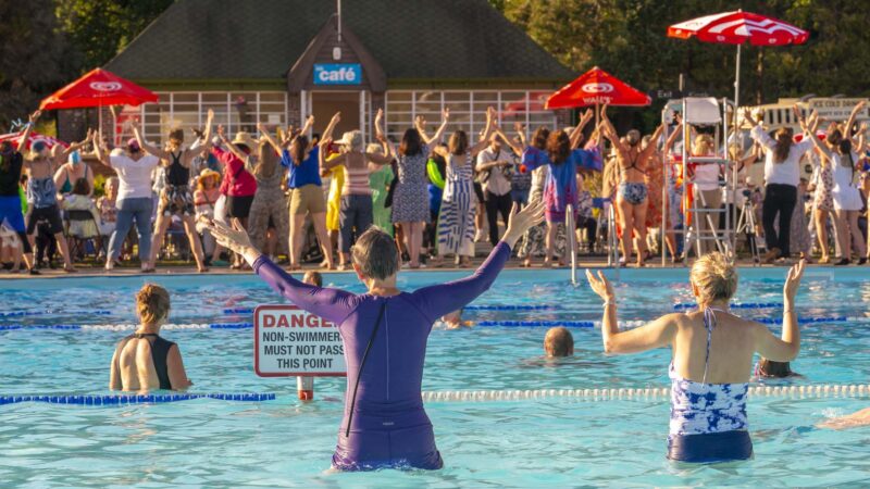 hundreds of people with arms in the air and 5 or 6 in a pool behind in background a cafe sign and sign in water danger non swimmers must not pass this point