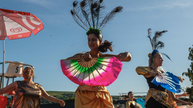 3 people with peacock feathers in head dress and holding colourful dance fans ilkley moors are in the back ground