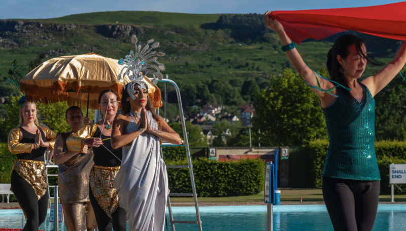 bollywood dancers in procession around a pool wearing colourful lavish indian costumes